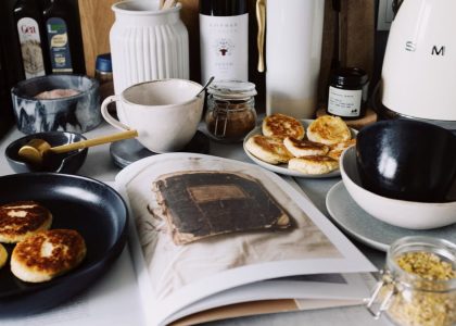 One portion of homemade cheese pancakes in frying pan and another on plate locating near opened stylish magazine among kitchen utensils and appliances in bright kitchen at home